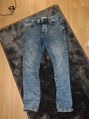 Blå slim fit jeans med tvättad look - Säljer ett par blå slim fit jeans med snyggt tvättad effekt och klassisk femficksdesign. Jeansen har normal midja, dragkedjegylf och bälteshällor. Perfekta för dig som gillar en avslappnad men trendig stil.Storlek W32 L32