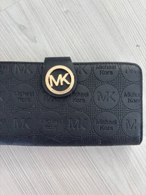 Svart Michael Kors plånbok - Snygg svart plånbok från Michael Kors med präglat MK-mönster över hela ytan och ett stort guldfärgat MK-emblem på framsidan. Plånboken är avlång och tillverkad i tåligt konstläder med struktur. Perfekt för dig som gillar stilrena accessoarer med logga.