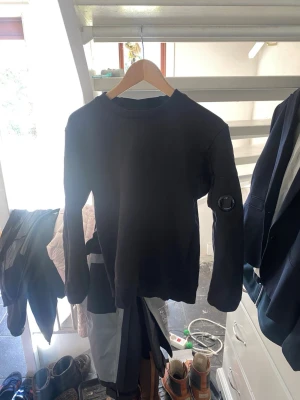 Svart sweatshirt från C.P. Company - Säljer en svart sweatshirt från C.P. Company med deras ikoniska linsdetalj på ärmen. Tröjan har rund halsringning och långa ärmar. Perfekt för dig som gillar streetwear och vill ha något stilrent men ändå unikt.