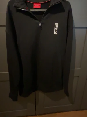 Svart halvzip tröja från HUGO - Svart långärmad tröja från HUGO med halvzip och hög krage. Tröjan har en vit HUGO-logga på bröstet och ribbade muddar vid ärmslut. Perfekt för en clean och stilren look.