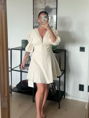 klänning från Adoore mer som strl 38 - Supersöt vit kortklänning från Adoore med puffärmar, v-ringning och knappar framtill. Klänningen har ett diskret broderat mönster och lätt utsvängd kjol. Perfekt för sommarens alla tillfällen!