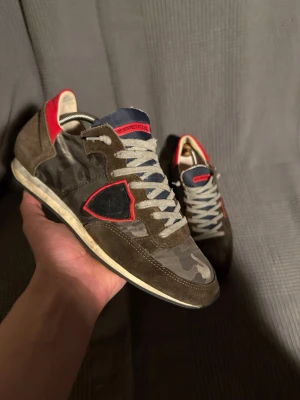 Philippe Model camo sneakers brun/röd - Snygga sneakers från Philippe Model i brun mocka och kamouflagemönstrad textil. Skorna har röda detaljer vid hälen och sidan, samt grå skosnören och mörkblå plös med röd logga. Klassisk låg modell med vit sula och coola kontraster.