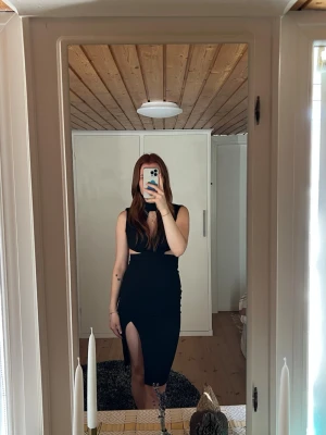 Svart långklänning med slits - Snygg svart långklänning med hög hals, ärmlös design och cut out-detaljer i midjan. Klänningen har en djup slits framtill och en figurnära passform som framhäver siluetten. Perfekt för dig som vill sticka ut med en modern och trendig look.