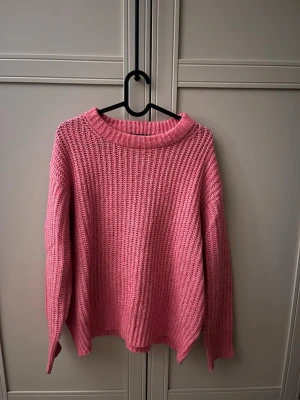 Rosa stickad tröja med grov struktur - En rosa stickad tröja med grov struktur och rund hals. Tröjan har långa ärmar och ribbade muddar vid hals, ärmslut och nederkant. Perfekt för att hålla sig varm och snygg under kyliga dagar. Storlek S och tröjan kommer från lager 157
