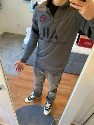 Grå PSG träningströja från Nike - Snygg grå långärmad träningströja från Nike med PSG-logga och svart tryck på bröstet. Tröjan har halv dragkedja framtill och röda detaljer på ärmen. Perfekt för fotbollsträning eller gymmet.