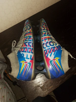 Nike Phantom GT2 färgglada fotbollsskor - Säljer ett par Nike Phantom GT2 fotbollsskor med färgglad grafik i blått, rött, gult och vitt. Skorna har snörning, textilovandel och syntetdetaljer. Perfekta för dig som vill sticka ut på planen med unika skor och grym passform.