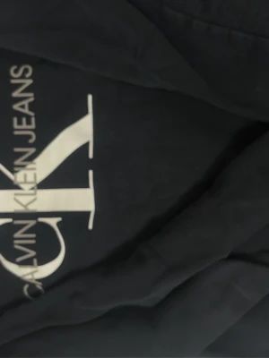 Svart hoodie från Calvin Klein Jeans - Snygg svart hoodie från Calvin Klein Jeans med stor vit CK-logga och text på bröstet. Tillverkad i mjukt och varmt material med borstad insida för extra komfort. Perfekt för dig som gillar klassisk streetwear och vill ha en hoodie med ikonisk logga.