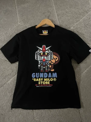 Svart Gundam x Baby Milo T-shirt BAPE - Säljer en svart t-shirt från A Bathing Ape med ett färgglatt Gundam x Baby Milo-tryck på bröstet. Klassisk rund hals och korta ärmar. Perfekt för dig som gillar streetwear och anime. Materialet är mjuk bomull.