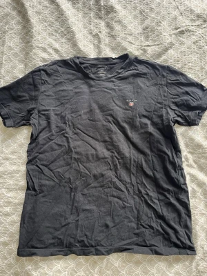 Svart t-shirt från Gant - En klassisk svart t-shirt från Gant med liten logga på bröstet. T-shirten har rund hals och korta ärmar, perfekt för en enkel och stilren look. Tillverkad i mjuk bomull som känns skön mot huden.