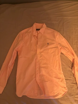 Ljusrosa skjorta från Ralph Lauren - Klassisk ljusrosa skjorta från Ralph Lauren med button-down krage och broderad logga på bröstet. Skjortan har långa ärmar och knäppning framtill. Perfekt för dig som gillar stilrena och tidlösa plagg.