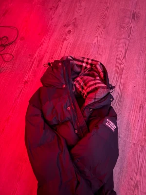  Burberry jacka/väst - Säljer en svart pufferjacka/väst från Burberry med klassiskt rutigt foder i huvan. Jackan har dragkedja och knappar framtill samt en hög krage. Perfekt för kalla vinterdagar och riktigt snygg med sin ikoniska design. Samt avtagbar armar så man kan göra den till bäst