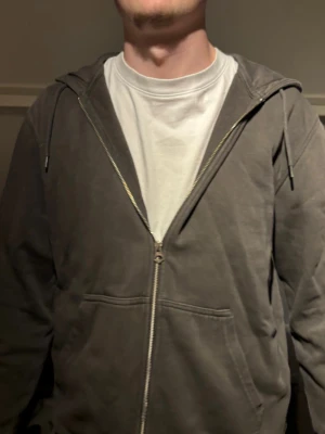 Grå zip hoodie med huva - En enkel och stilren grå hoodie med dragkedja framtill och två fickor. Hoodien har en klassisk huva med dragsko och ribbade muddar vid ärmslut och nederkant. Perfekt för en avslappnad och chill look.