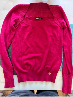 Bår Mörkrosa tröja  - Mysig mörkrosa  tröja  med bred ribbad krage och långa ärmar. Tröjan har en enkel, stilren design och en liten metallbricka vid nederkanten. Perfekt för dig som gillar färg och vill ha något bekvämt.