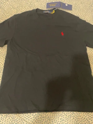 Svart t-shirt från Polo Ralph Lauren - Klassisk svart t-shirt från Polo Ralph Lauren med röd broderad logga på bröstet. T-shirten har rund halsringning och korta ärmar. Tillverkad i mjuk bomull som känns skön mot huden. Perfekt basplagg med stilren design.