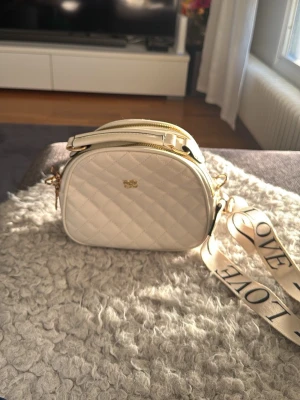 Vit quiltad axelväska med bred rem - Snygg vit axelväska i quiltat mönster med guldfärgade detaljer och dragkedja. Väskan har en bred axelrem med texten 'LOVE' i svart, samt en liten ficka med dragkedja på baksidan. Perfekt för dig som vill ha en trendig och praktisk accessoar. Knappt använd. 23x20x9 cm