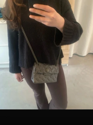 Grå quiltad handväska Kurt Geiger - Snygg grå quiltad handväska från Kurt Geiger med kedjerem och lock med magnetknäppning. Väskan har ett elegant mönster och metallogo framtill. Perfekt storlek för det viktigaste och levereras med original dustbag. Liten defekt i hörnet av väskan men märks inte. 