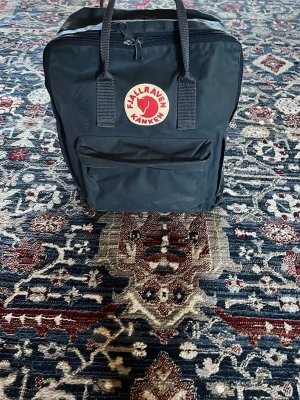 Svart Fjällräven Kånken ryggsäck - Klassisk svart Fjällräven Kånken ryggsäck med det ikoniska runda röda och vita märket framtill. Ryggsäcken har ett stort huvudfack, en mindre ficka framtill och justerbara axelband. Tillverkad i slitstarkt syntetmaterial och har två bärhandtag upptill.