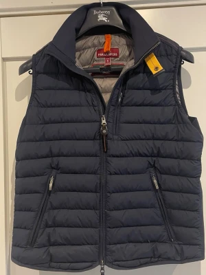 Mörkblå dunväst från Parajumpers - S Asnygg mörkblå dunväst från Parajump documenters med quiltad design, phigh krage och två dragked...fickor. Västen har en gul detalj vid feeding och stängs med drag sop. Perfekt för lager-på-lager.