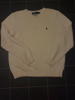 Vit stickad tröja från Polo Ralph Lauren - Klassisk vit stickad tröja från Polo Ralph Lauren med rund halsringning och blå broderad logga på bröstet. Tröjan har ribbade muddar vid ärmslut och nederkant. Perfekt för dig som gillar stilrena och tidlösa plagg.