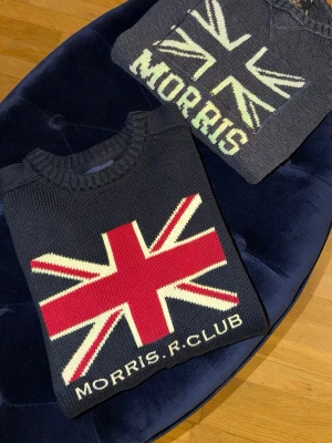 Stickad tröja Morris R. Club - Snygg stickad tröja från Morris R. Club med stor Union Jack-flagga framtill. Tröjan är mörkblå med röd, vit och beige design och har ribbade muddar vid hals, ärmslut och nederkant. Perfekt för dig som gillar preppy stil och vill sticka ut. Den med röd är storlek M den andra S