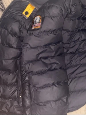 Svart pufferjacka från Parajumpers - Snygg svart pufferjacka från Parajumpers med klassisk patch på bröstet och gul detalj vid kragen. Jackan har quiltad design, dragkedja framtill och är tillverkad i ett glansigt material som ger en modern look. Perfekt för kalla vinterdagar.