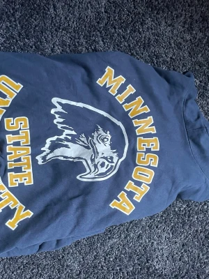 Blå Minnesota State University hoodie - Snygg mörkblå hoodie med tryck från Minnesota State University och en stor örn på bröstet. Gula och vita bokstäver ger en klassisk collegekänsla. Perfekt för dig som gillar sportig och avslappnad stil. Mjuk bomullskänsla och huva för extra komfort.