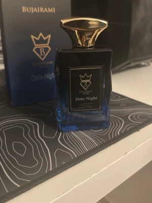 Bujairami Date Night parfym 100ml - Exklusiv parfym från Bujairami, Date Night, i en elegant mörkblå glasflaska med guldfärgat lock och detaljer. Flaskan rymmer 100 ml och har en lyxig design med svart och guld etikett. Perfekt för dig som vill ha en unik och sofistikerad doft.