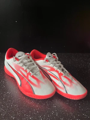 Puma vita och röda fotbollsskor - Snygga fotbollsskor från Puma i vitt och rött med svarta detaljer. Skorna har en låg profil, snörning och mönstrad sula för bra grepp på inomhusplan. Ovandelen är i syntetmaterial och insidan är fodrad i rött. Perfekta för dig som vill sticka ut på planen.