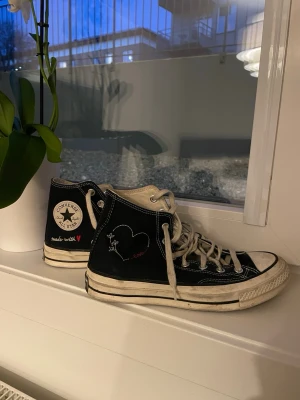 Converse All Star svart hjärtdesign - Säljer ett par svarta Converse All Star high tops med vit sula och snörning. Skorna har en unik hjärtdesign med texten 'made with ♥' på sidan och en röd hjärtdetalj under sulan. Klassisk rund tå och canvasmaterial.