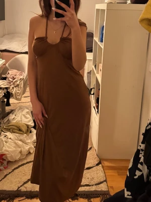 Brun långklänning med smala axelband - Säljer en enkel och stilren brun långklänning med smala axelband och figurnära passform. Klänningen har en mjuk, följsam känsla och en lätt v-ringad urringning. Perfekt för dig som gillar minimalistisk stil.