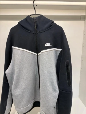 Nike tech fleece - Nike hoodie i svart och ljusgrått med hel dragkedja framtill och vit logga på bröstet. Hoodien har huva, fickor på sidorna och en extra ficka med dragkedja på ärmen. Tillverkad i mjukt sweatshirtmaterial, perfekt för en avslappnad streetwear-look.