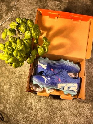 Blåa Puma fotbollsskor med snörning - Säljer ett par snygga blåa fotbollsskor från Puma med vita detaljer och logga på sidan. Skorna har snörning och är tillverkade i syntetmaterial för bästa grepp och känsla på planen. Perfekta för dig som vill sticka ut på fotbollsplanen!