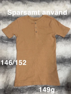 Brun ribbad t-shirt med knappar - Snygg beige t-shirt med ribbad struktur och tre knappar framtill. Kortärmad modell med rund halsringning, perfekt för en avslappnad stil. Passar dig som gillar enkla men stilrena plagg.🌸 Sparsamt använd. Storlek 146/152 