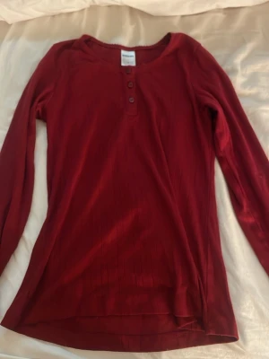 Vinröd långärmad topp med knappar - Vinröd långärmad topp med rund halsringning och tre knappar framtill. Tunn och mjuk kvalitet, perfekt för lager på lager. Enkel och stilren design som passar till mycket.