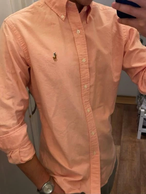 Rosa skjorta från Ralph Lauren (Slim Fit) Oxford - Klassisk persikofärgad skjorta från polo Ralph Lauren med button-down krage och broderad logga på bröstet. Skjortan har långa ärmar, vita knappar och är tillverkad i mjuk bomull. Perfekt för en stilren och avslappnad look. Storlek Small. Snabb Leverans!