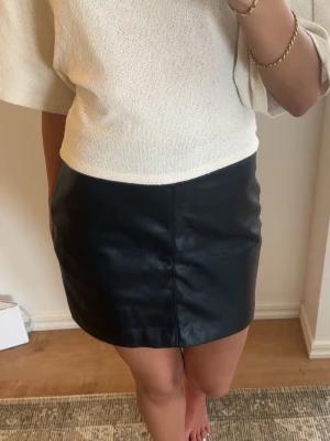 Svart kort skinnkjol med dragkedja - Snygg svart kort kjol i skinnimitation med synlig dragkedja bak. Kjolen har en enkel och stilren design som passar perfekt till många olika outfits. Materialet ger en cool känsla och kjolen har en normal passform.