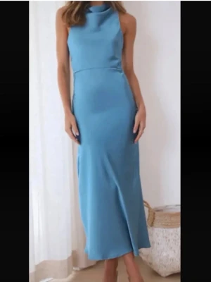 Blå långklänning från Hello Molly med öppen rygg - Snygg blå långklänning i satin med hög hals och korsade axelband i ryggen som ger en öppen rygg. Klänningen har en elegant, figurnära passform och släpper lätt från höften och ner. Perfekt för dig som vill sticka ut med en stilren look.