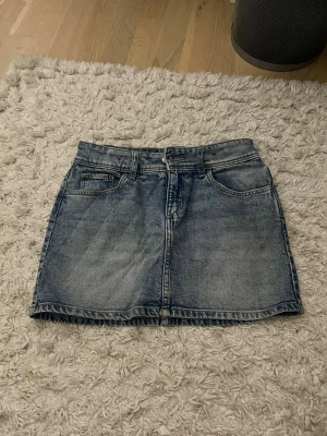 Blå jeanskjol  - Säljer en blå jeanskjol med fem fickor och klassisk knapp- och dragkedjestängning. Kjolen har en rak passform och är i kort modell, perfekt till vardagslooken. Passar till det mesta och är enkel att styla.