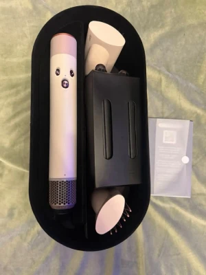 Dyson Airwrap multistyler i svart etui - Säljer en Dyson Airwrap multistyler med flera tillbehör i ett svart, ovalt etui. Verktyget är i ljusrosa och silverfärgat material och har flera utbytbara stylinghuvuden för hår. Perfekt för att skapa olika frisyrer med värme och luft.