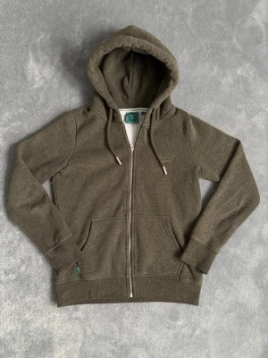 Olivgrön Superdry Hoodie  - Superdry hoodie. Endast använd ett fåtal gånger, nyskick. ”Superdry” loggan är broderad på vänstra bröst. Helt perfekt nu till våren och sommar kvällar för en clean look! Storlek S. Hör av dig vid minsta fundering!😊