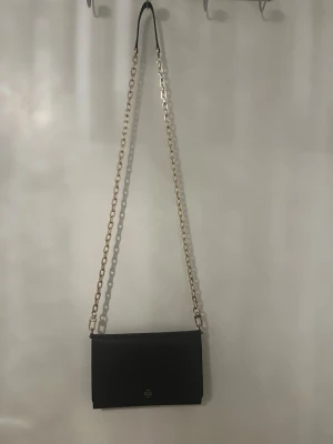 Svart kuvertväska från Tory Burch - Snygg svart kuvertväska från Tory Burch med guldfärgad kedjerem och diskret logga framtill. Väskan har en stilren, rektangulär form och är tillverkad i slätt läder. Perfekt att bära crossbody med den långa kedjan. Kedjan är avtagbar. Har endast använts ett fåtal gånger och är i ett mycket fint skick.