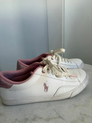 Rosa Ralph lauren skor - Snygga vita sneakers från Polo Ralph Lauren med rosa detaljer och logga på sidan. Skorna har rosa foder, glitterdetalj på hälen och klassiska vita snören. Dem är använda cirka 5-10 gånger. Pris kan diskuteras😊
