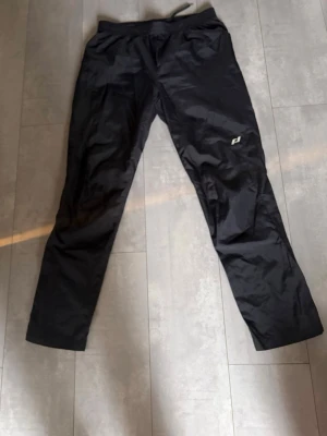 Svarta vindbyxot från Pro Touch - Säljer ett par svarta vind- och regnbyxor  från Pro Touch med resår i midjan och diskret logga på ena benet. Byxorna är tillverkade i ett lätt och glansigt material som passar perfekt för sport eller chill. Raka ben och enkel design.