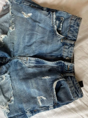 Blå denimshorts med slitningar - Säljer ett par blå denimshorts med råa kanter och slitna detaljer. 