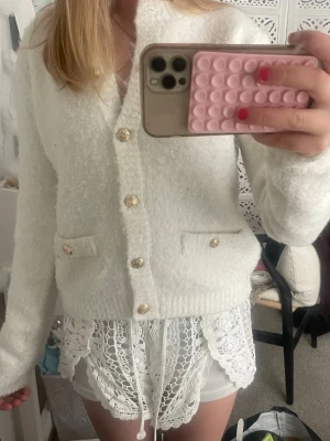 Vit fluffig kofta med guldknappar - Mysig vit kofta med fluffig textur och dekorativa guldfärgade knappar framtill. Koftan har två små fickor fram och ribbade muddar vid ärmslut och nederkant. Perfekt för lager på lager och ger en elegant touch till din outfit.