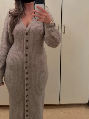 Beige ribbad lång stickad klänning - Lång stickad klänning i beige med ribbad struktur och v-ringning. Klänningen har långa ärmar och knappar hela vägen framtill, vilket ger en snygg och trendig look. Perfekt för dig som gillar en bekväm men ändå stilren outfit. nypris är 499kr