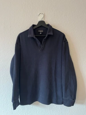Mörkblå rugbytröja från H&M - Klassisk mörkblå rugbytröja från H&M med krage och v-ringad hals. Tröjan har långa ärmar och ribbade muddar vid ärmsluten. Tillverkad i mjukt bomullsmaterial som ger en avslappnad och stilren look. 