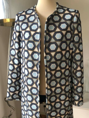 Mönstrad blå och beige kappa - Unik kappa med grafiskt hexagonmönster i blått, beige och vitt. Rak modell med rundad halsringning och dolda fickor i sidorna. Tjockare tyg med struktur som ger en cool retrovibe. Perfekt för dig som vill sticka ut med din stil.