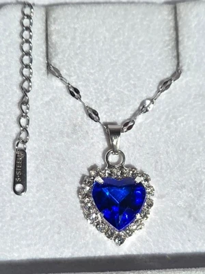 Halsband med blå hjärtformad sten - Snyggt halsband i rostfritt stål med en stor, blå hjärtformad sten som hänge. Runt stenen sitter gnistrande klara stenar som ger extra bling. Kedjan har ett elegant mönster och justerbar längd. Perfekt accessoar för dig som vill sticka ut.