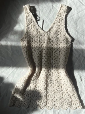 Beige spetslinne med v-ringning - Säljer ett superfint beige spetslinne med v-ringning och ärmlös design. Linnet har ett vackert blommigt spetsmönster och vågig kant nedtill. Perfekt för dig som gillar romantisk och feminin stil.